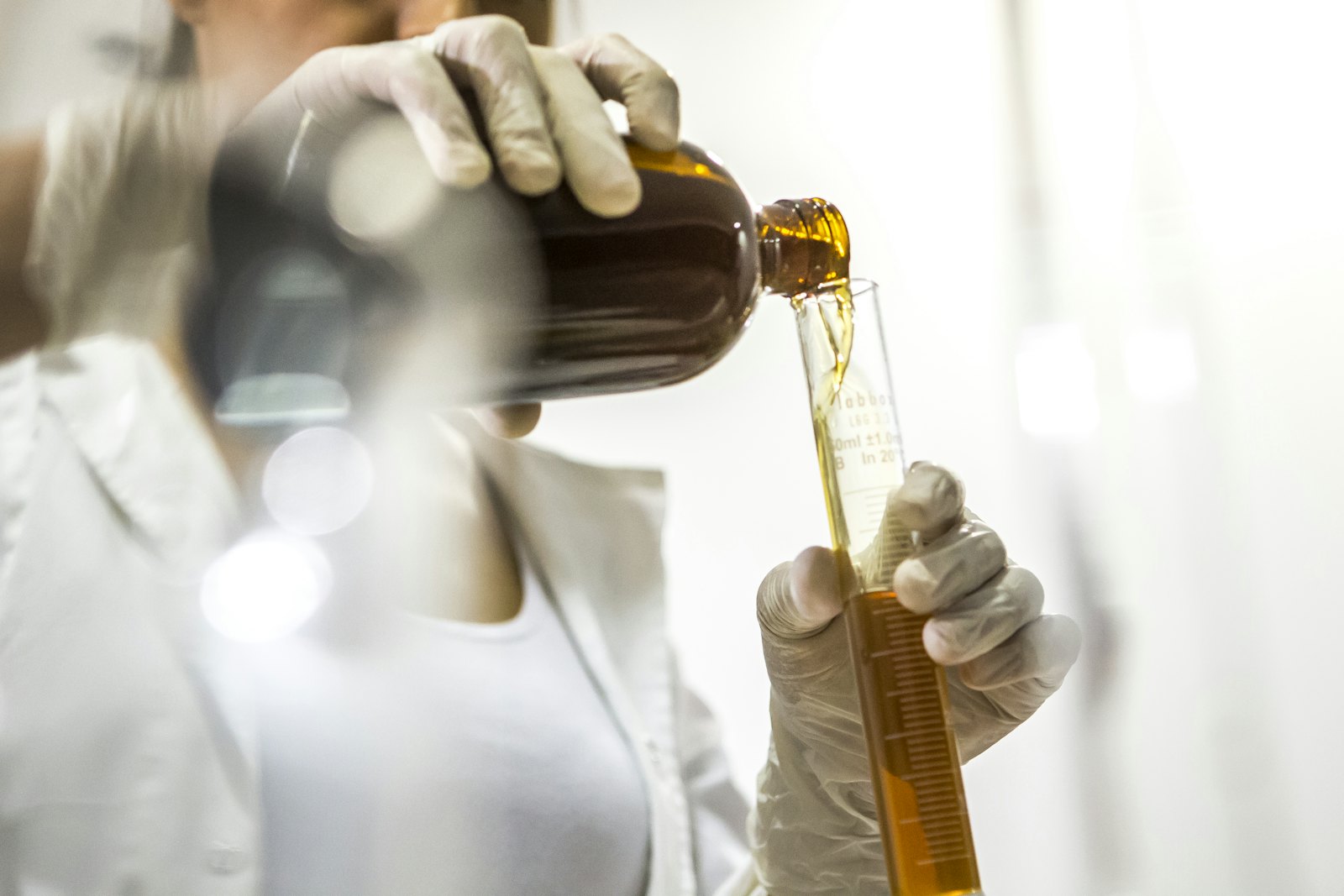 A gloved hand measuring an amber oil into a graduated cylinder